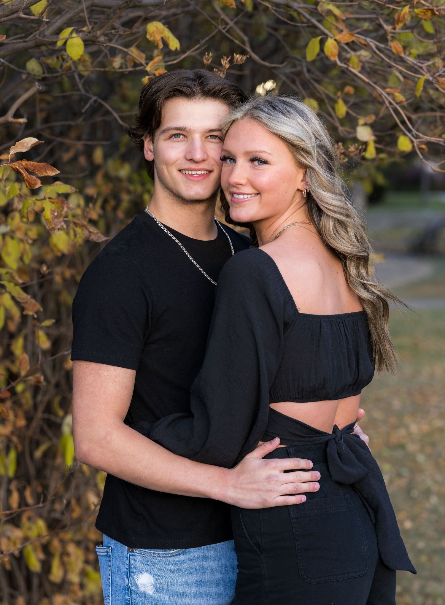 Stylish couple in black outfits posing against autumn foliage backdrop.
