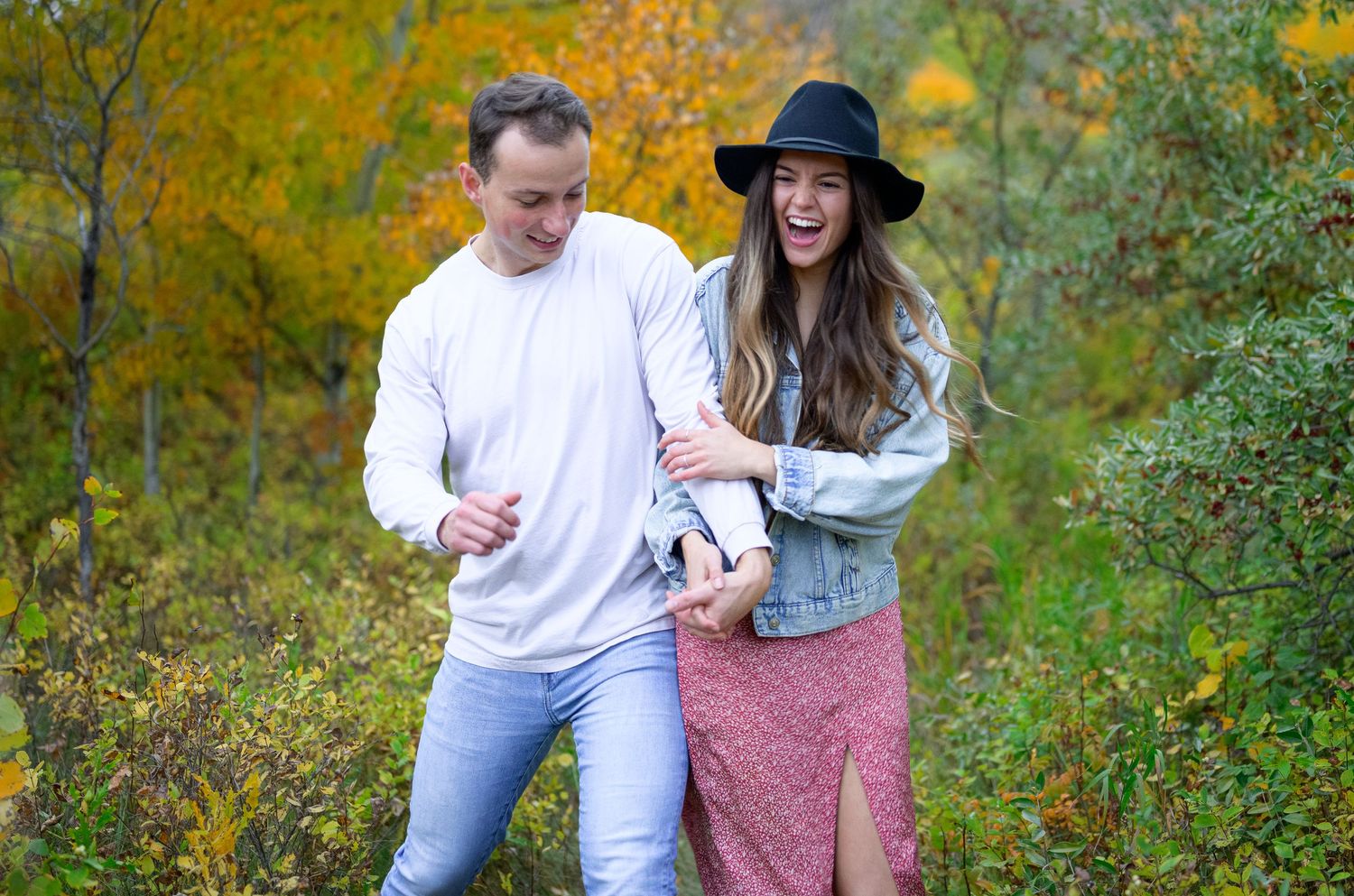 A couple walks together through autumn foliage wearing casual attire.