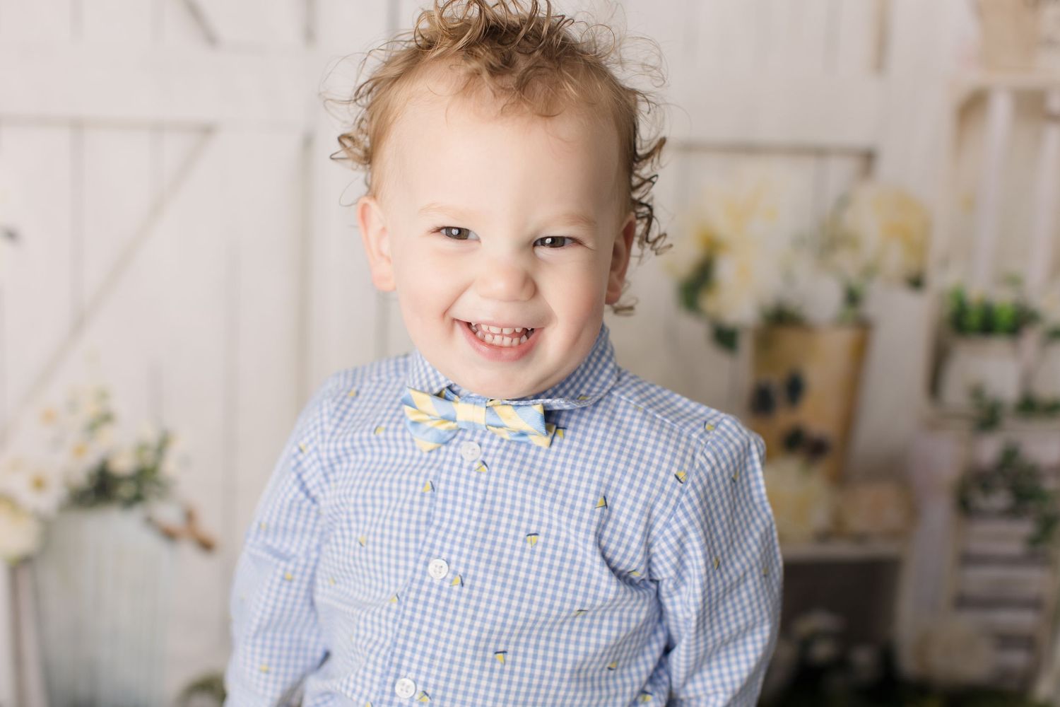 Young child in blue gingham shirt smiles brightly against white backdrop with floral decorations.