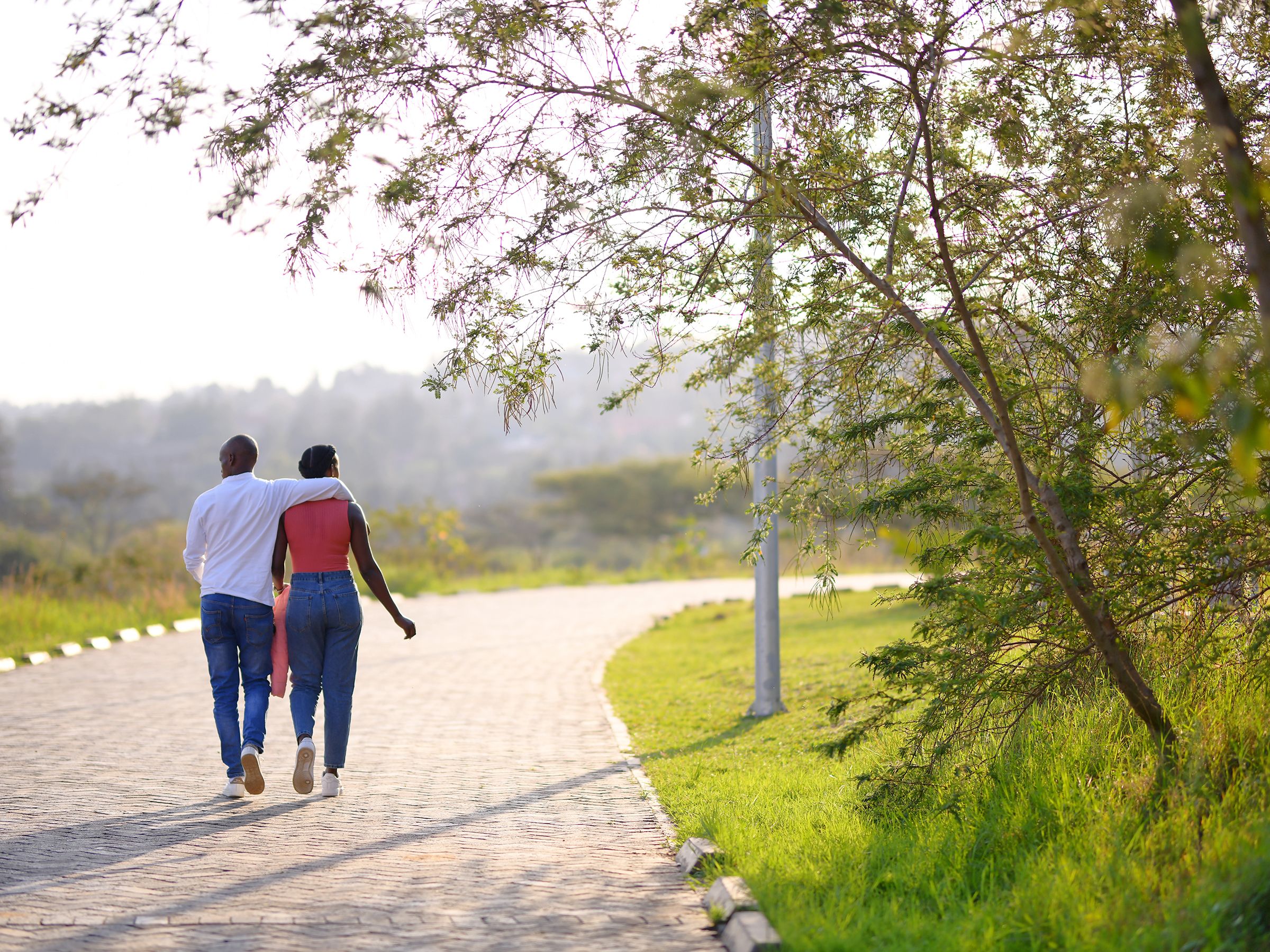 A couple walking together on a paved path surrounded by green trees and grass on a sunny day.