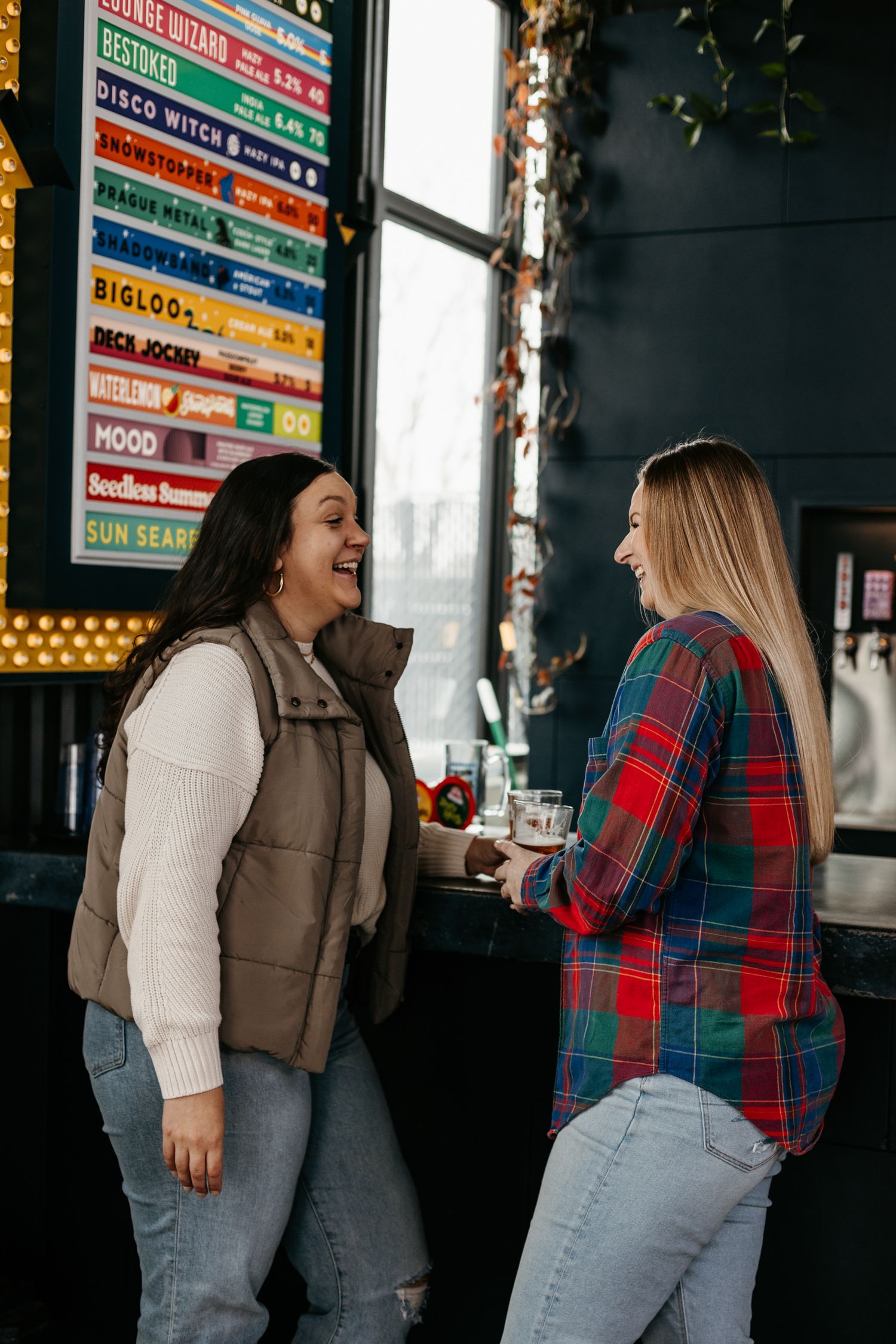 Two people laughing and socializing by a vintage menu board in an industrial-style cafe.