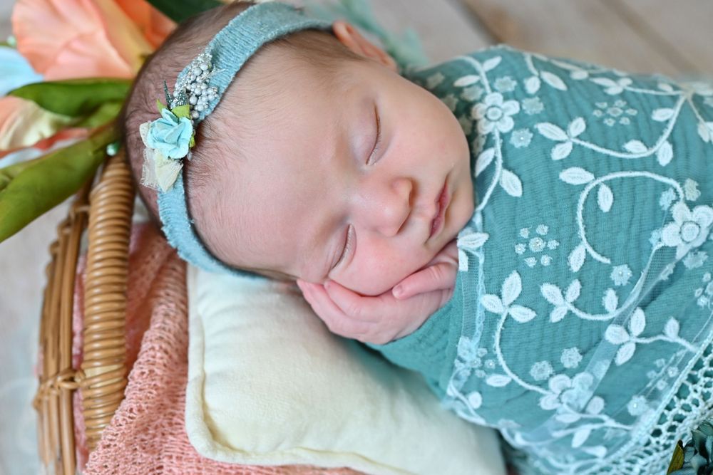 Sleeping newborn baby wrapped in teal floral swaddle with matching headband resting in a wicker basket.