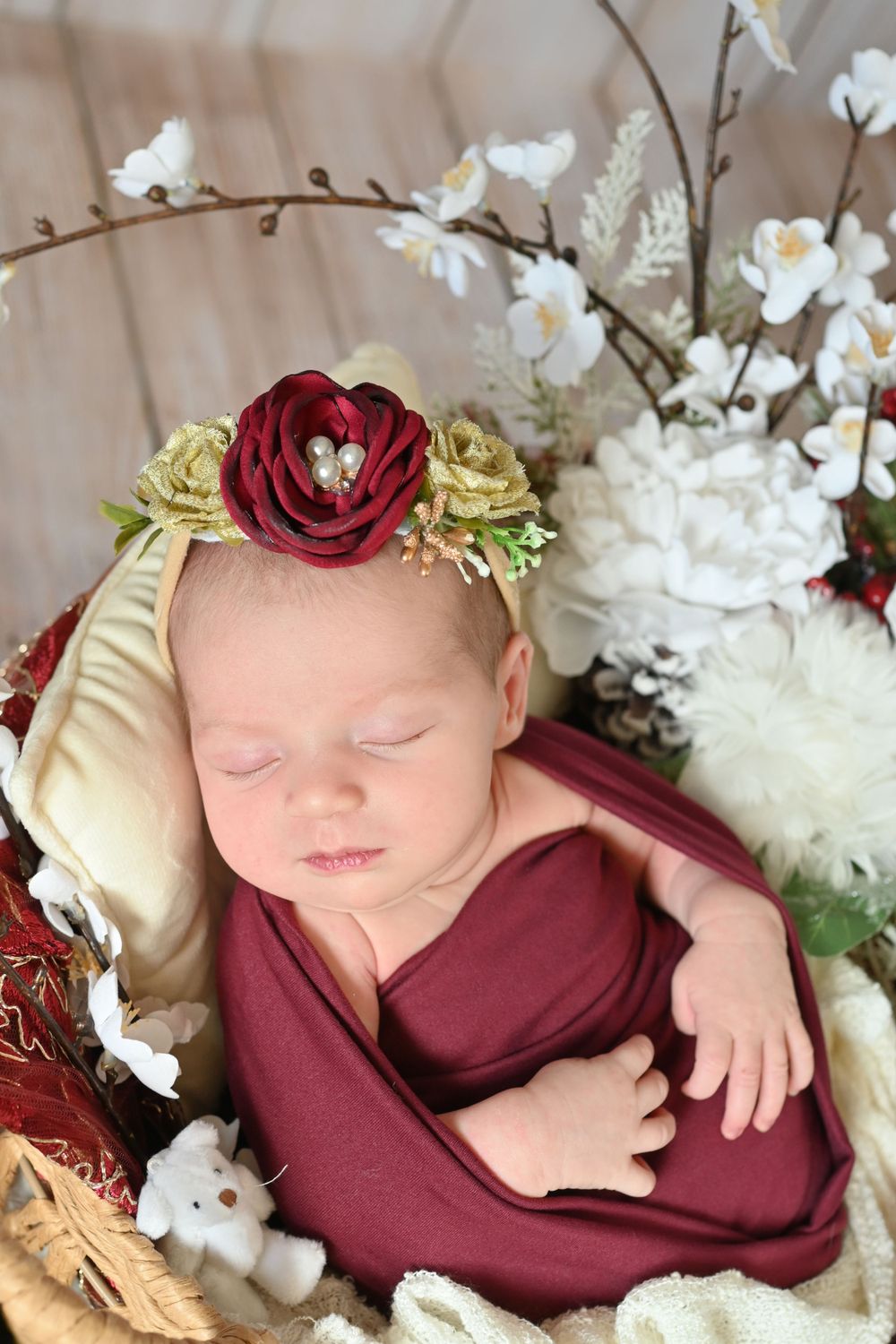 Sleeping infant wrapped in burgundy fabric with floral crown and white flowers.