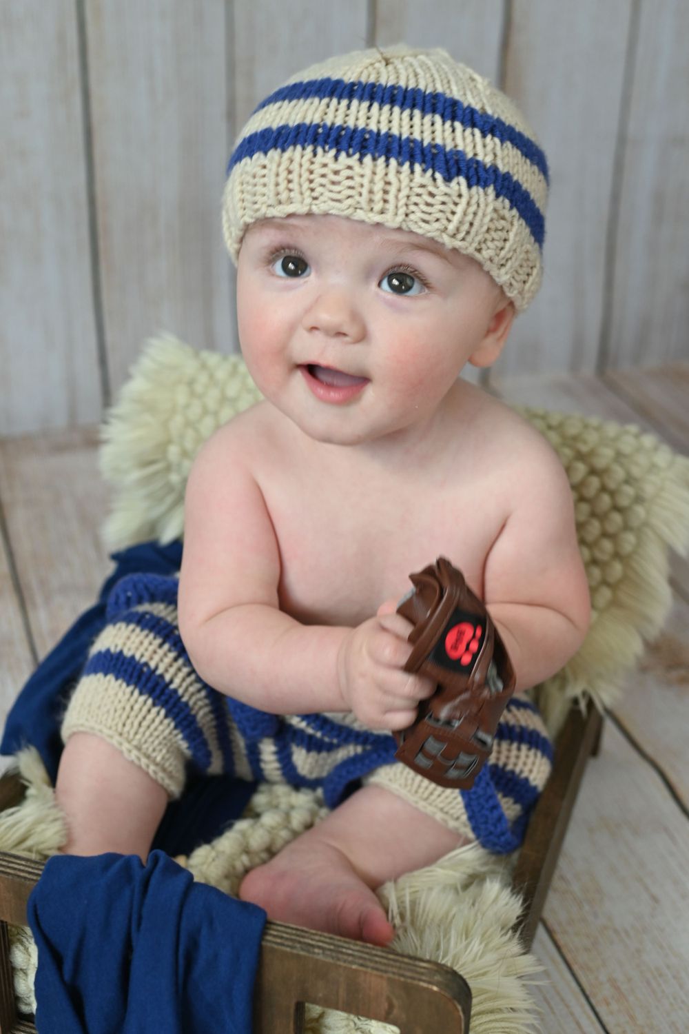 Smiling baby wearing striped knit hat and legwarmers holds toy baseball glove.