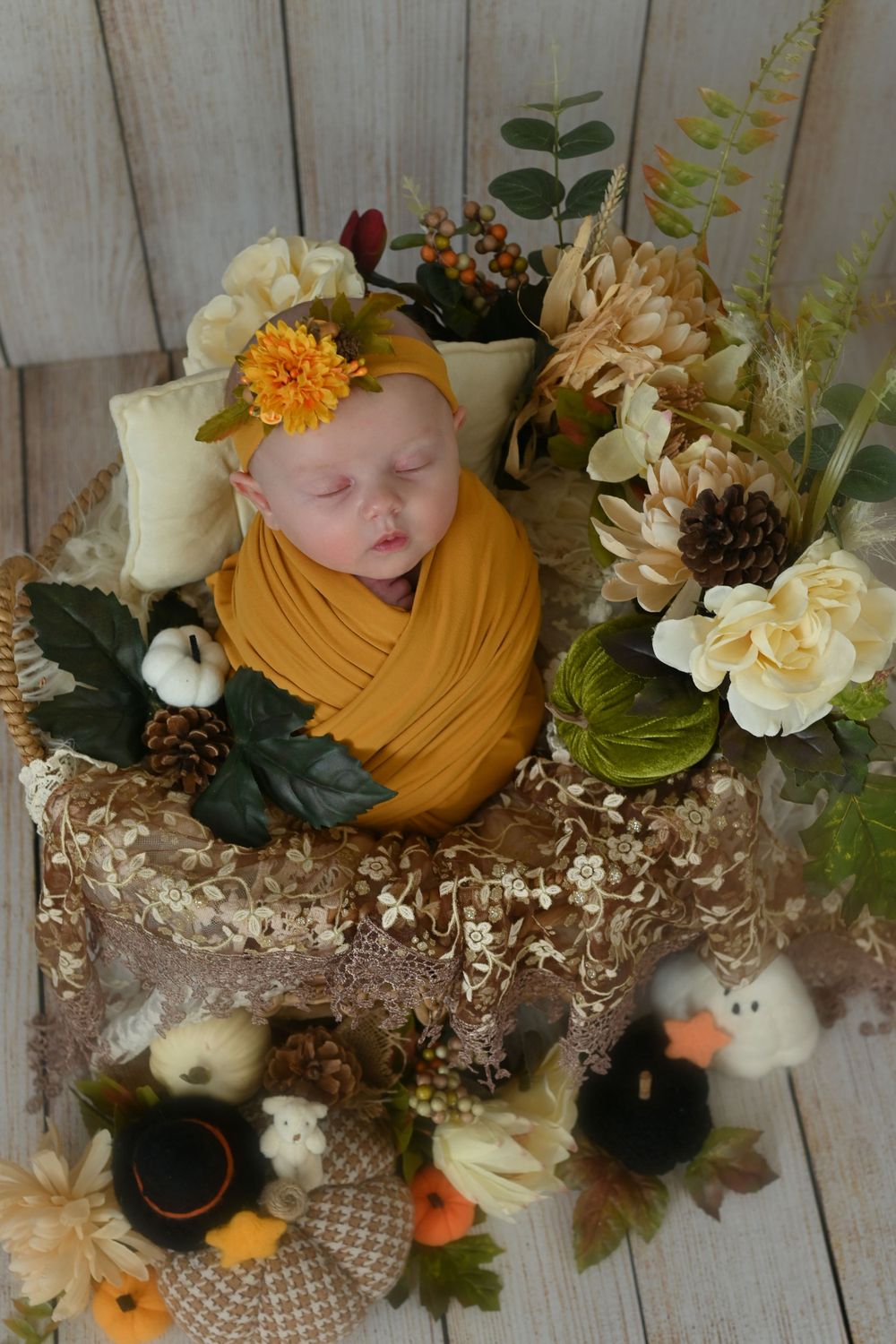 Sleeping newborn wrapped in mustard fabric surrounded by autumn flowers and pinecones.