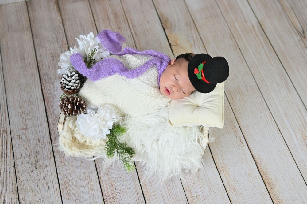 Newborn baby sleeping in a wooden bowl wearing a black pilgrim hat, wrapped in white, with pine cones and flowers.