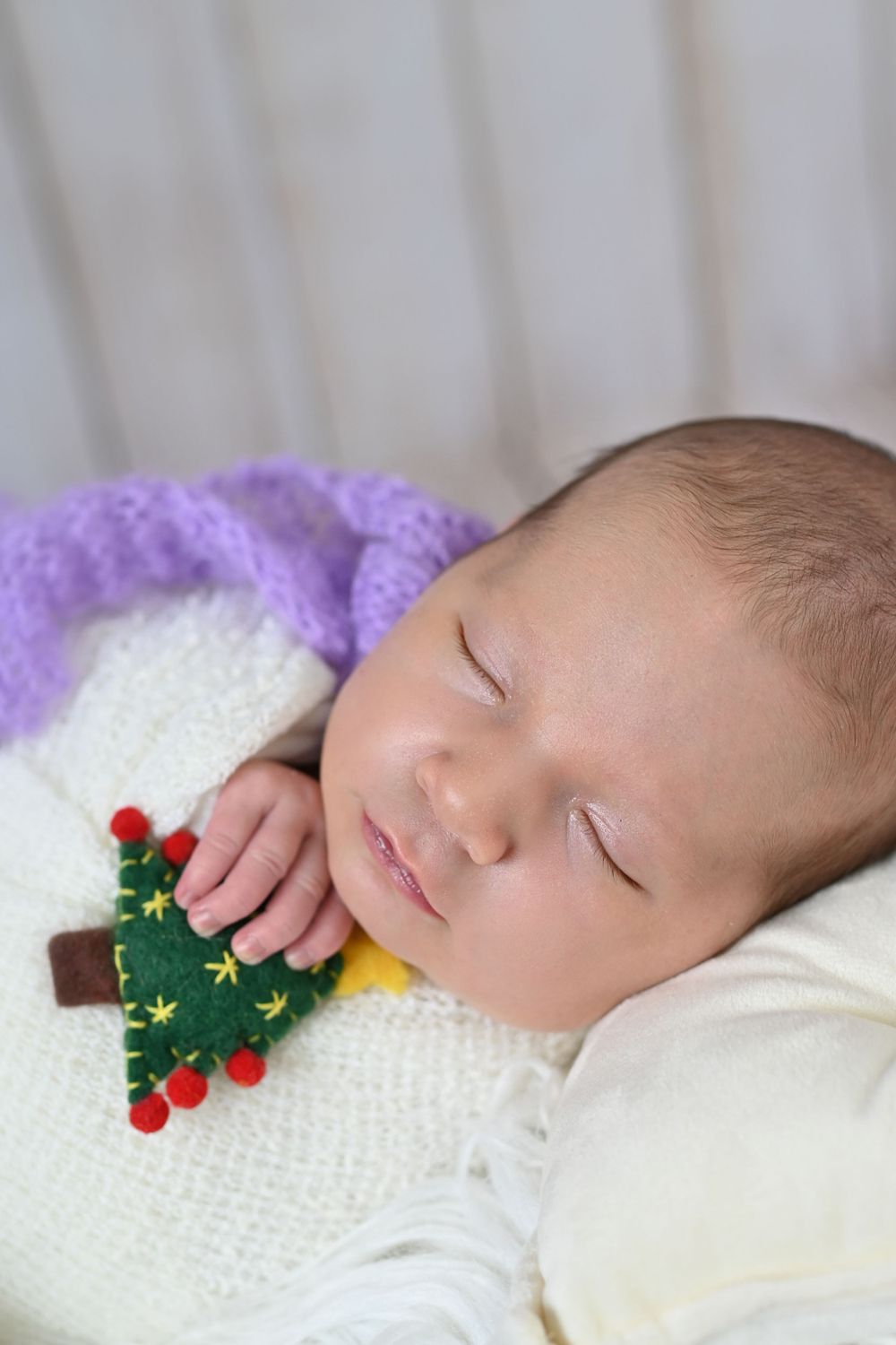 Sleeping newborn baby wrapped in white knit blanket with purple accent, holding a small felt Christmas tree ornament.