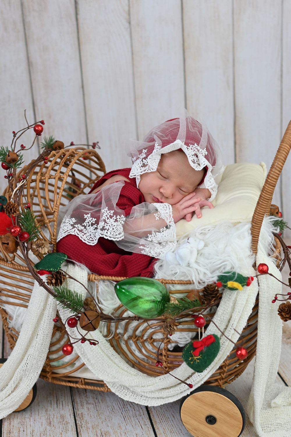 Sleeping infant in lace bonnet rests in wicker carriage with holiday decorations.