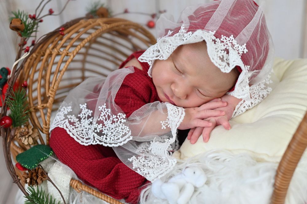Sleeping newborn baby in red outfit with white lace bonnet in wicker chair with Christmas decorations.