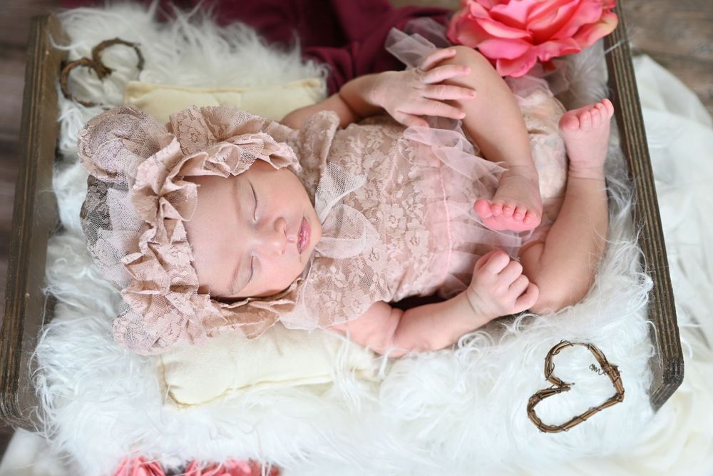 Sleeping newborn baby in pink lace outfit and headband on fluffy white fur blanket in wooden box.