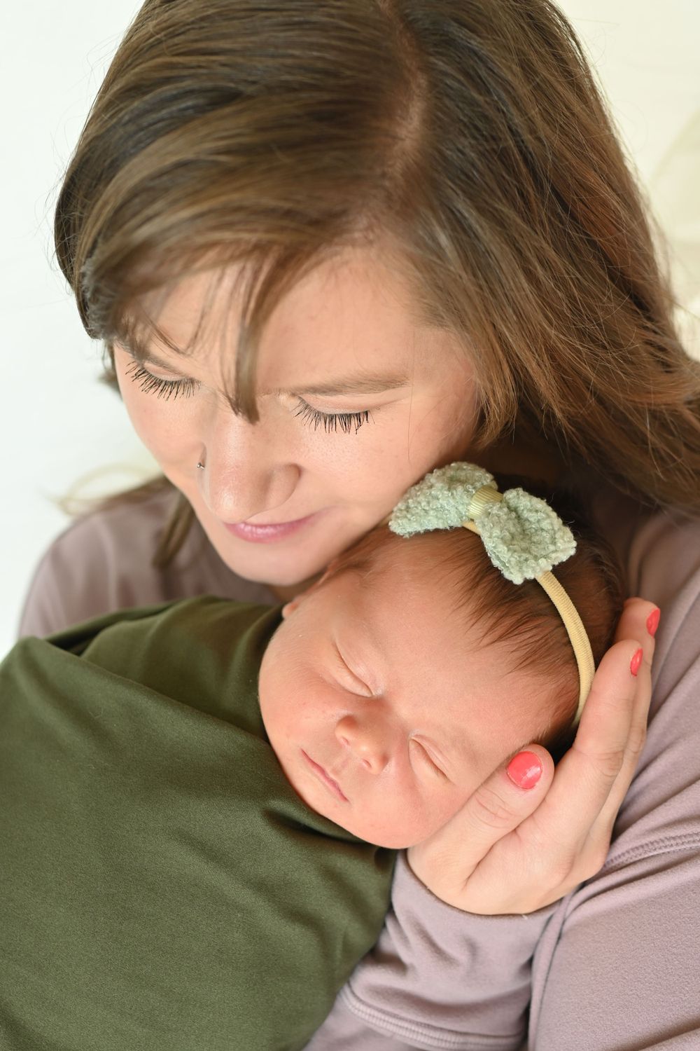 A mother tenderly holds her sleeping newborn baby girl wearing a green bow headband against her chest.