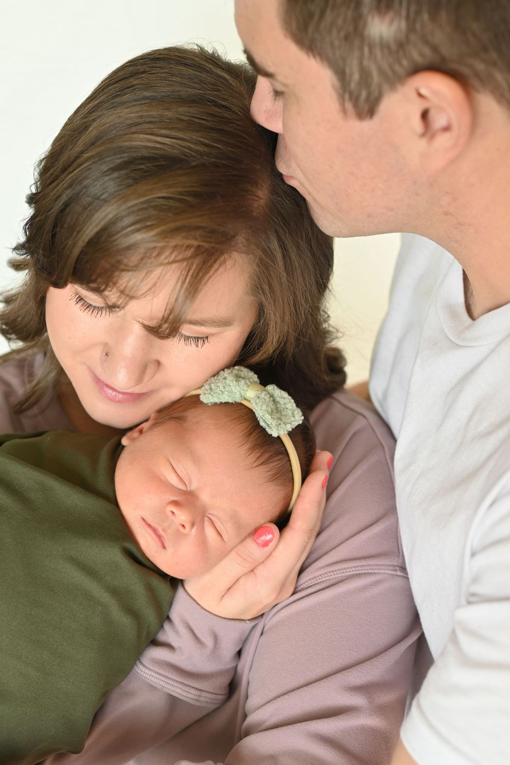 Parents lovingly gaze at their sleeping newborn baby girl wearing a green bow headband, wrapped in a soft blanket.