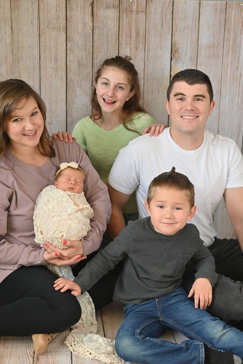 Family portrait with newborn baby in cream dress against wooden wall backdrop.