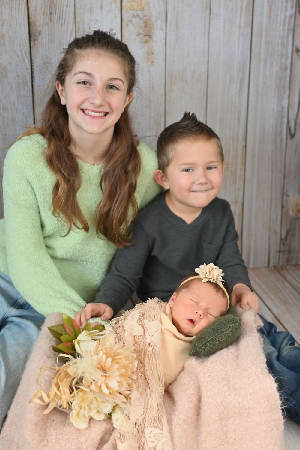 Sleeping newborn in cream lace outfit with siblings against rustic wooden backdrop.