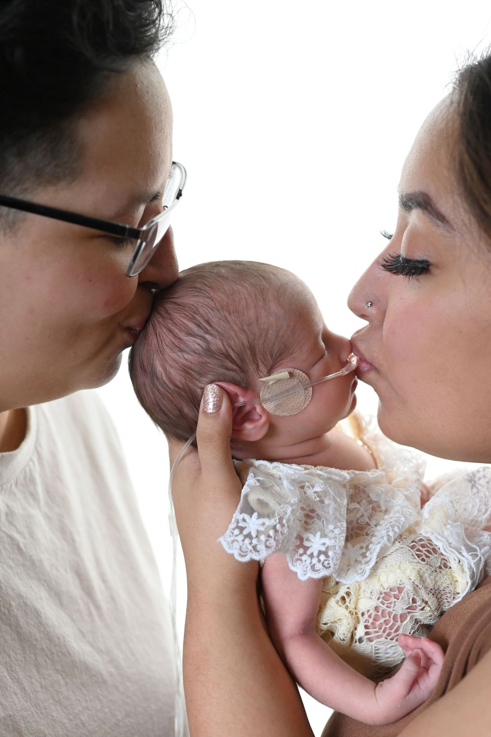 Tender moment with newborn baby in white lace dress against white background.