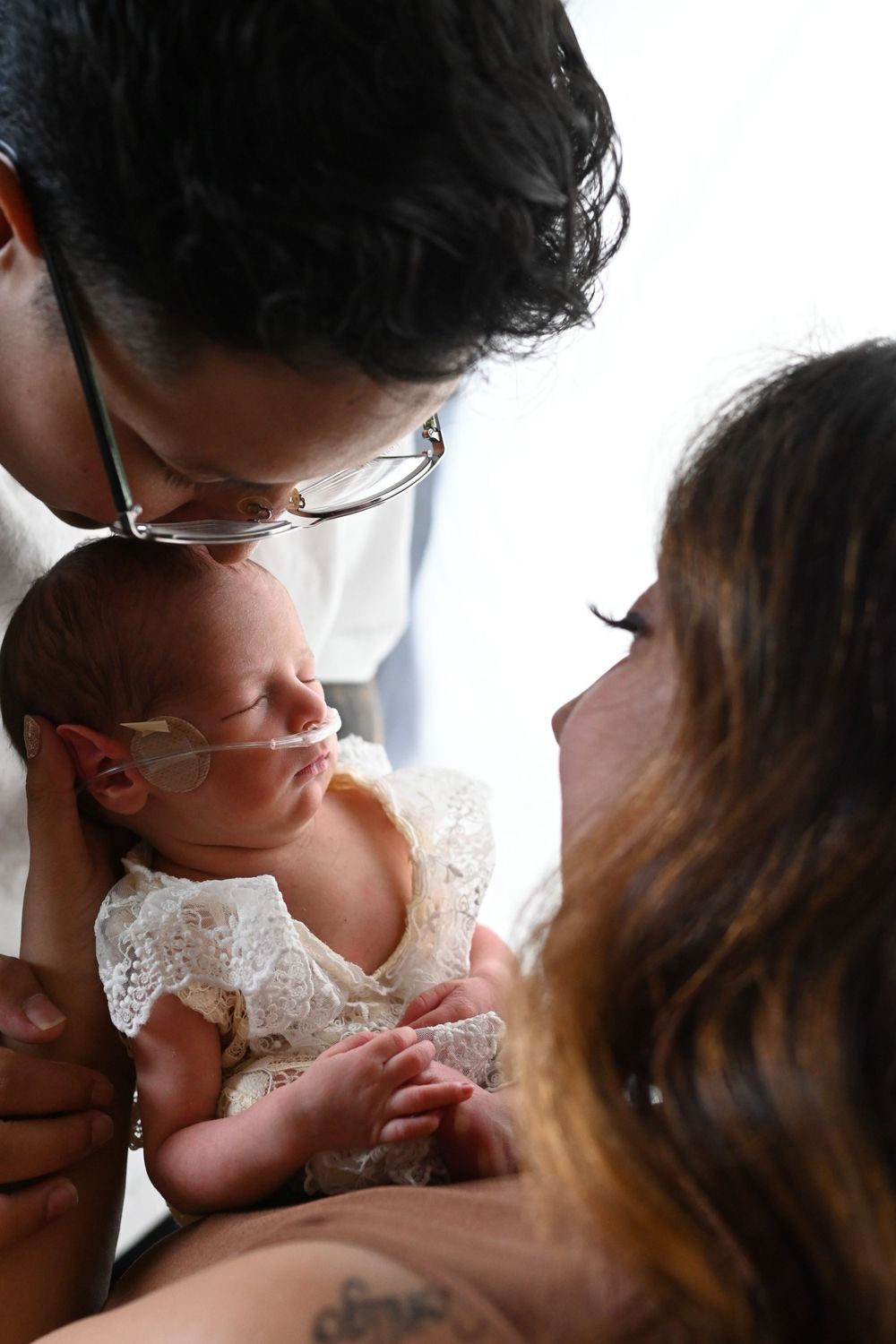 Intimate family portrait with newborn baby in lace dress against bright background.
