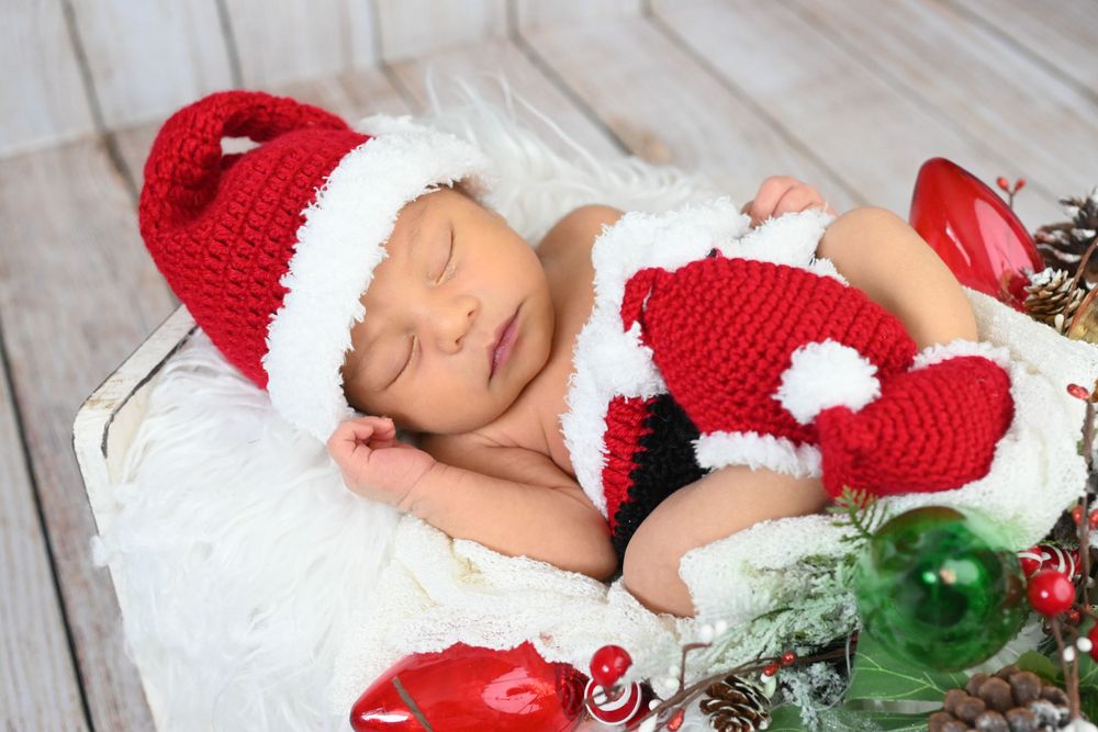 Newborn wearing red and white Santa hat sleeping among Christmas decorations.