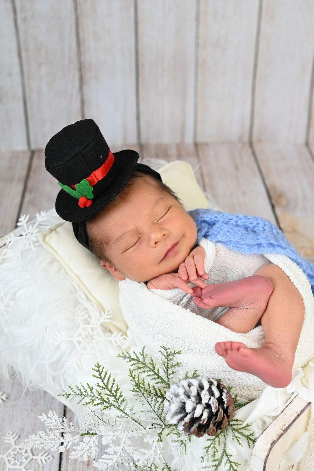 Sleeping baby with tiny black top hat posed with winter pinecones and greenery.