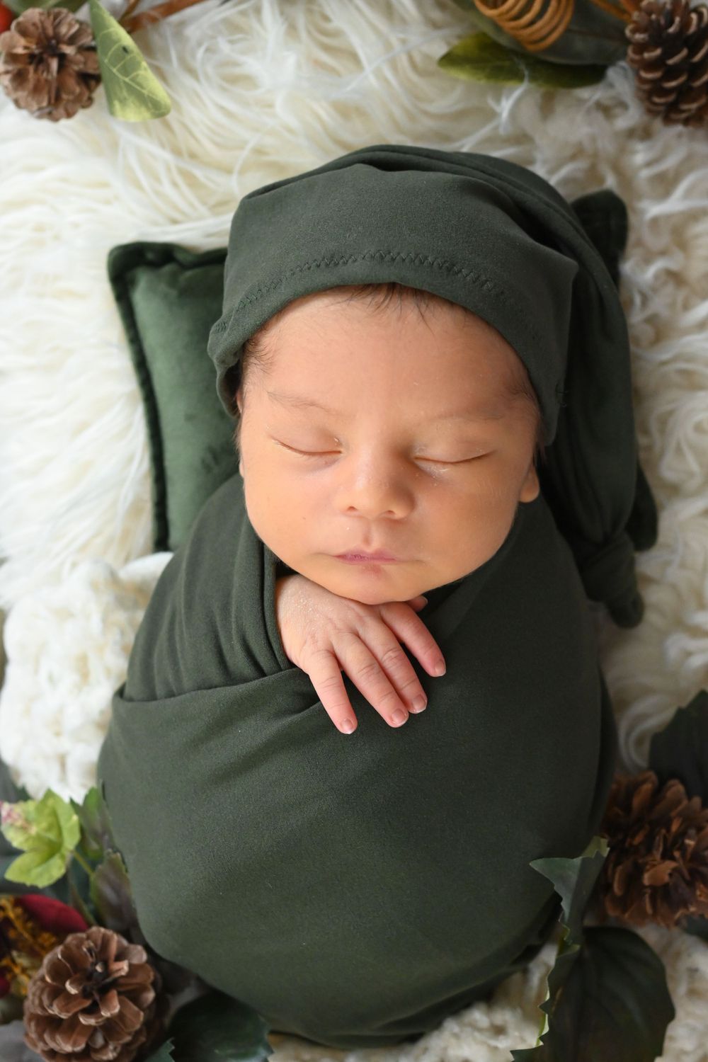 Newborn in dark green wrap sleeping peacefully with pinecones and holiday decorations.