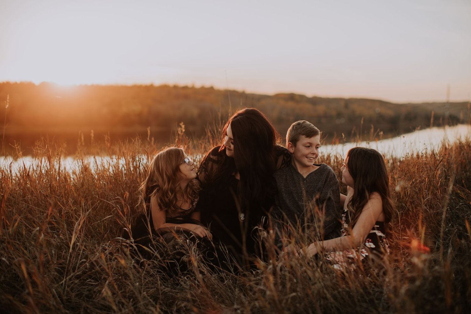 Silhouetted group sitting together in tall grass at sunset near a lake.