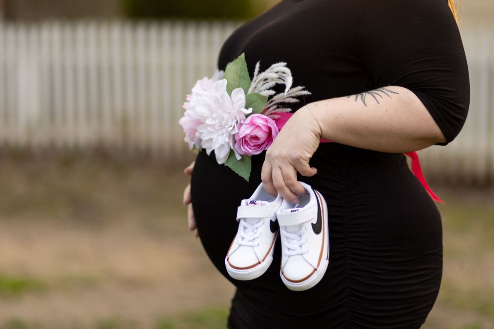 A pregnant woman in black dress holds tiny white baby shoes with pink floral accessories.