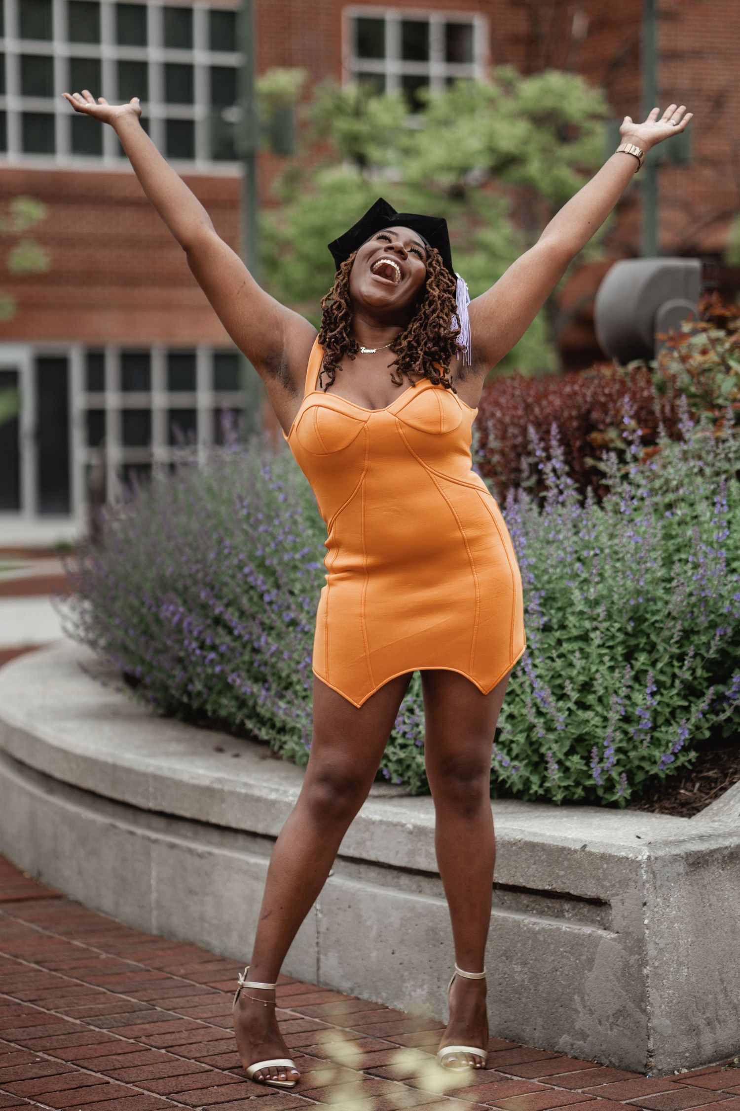 Graduate in orange dress celebrating with arms raised outdoors.