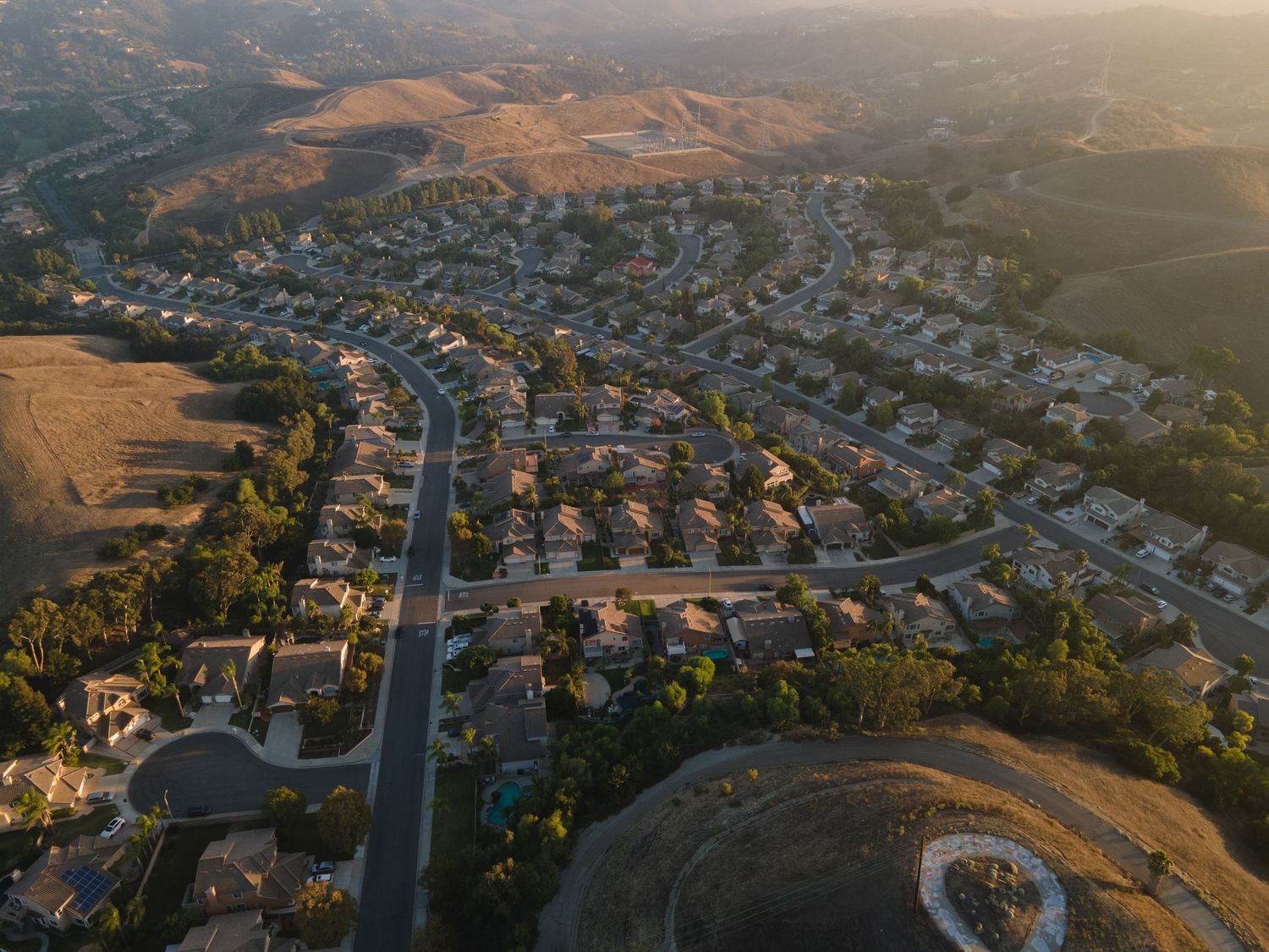 Aerial view of a sprawling suburban neighborhood nestled between golden hills in warm evening sunlight.