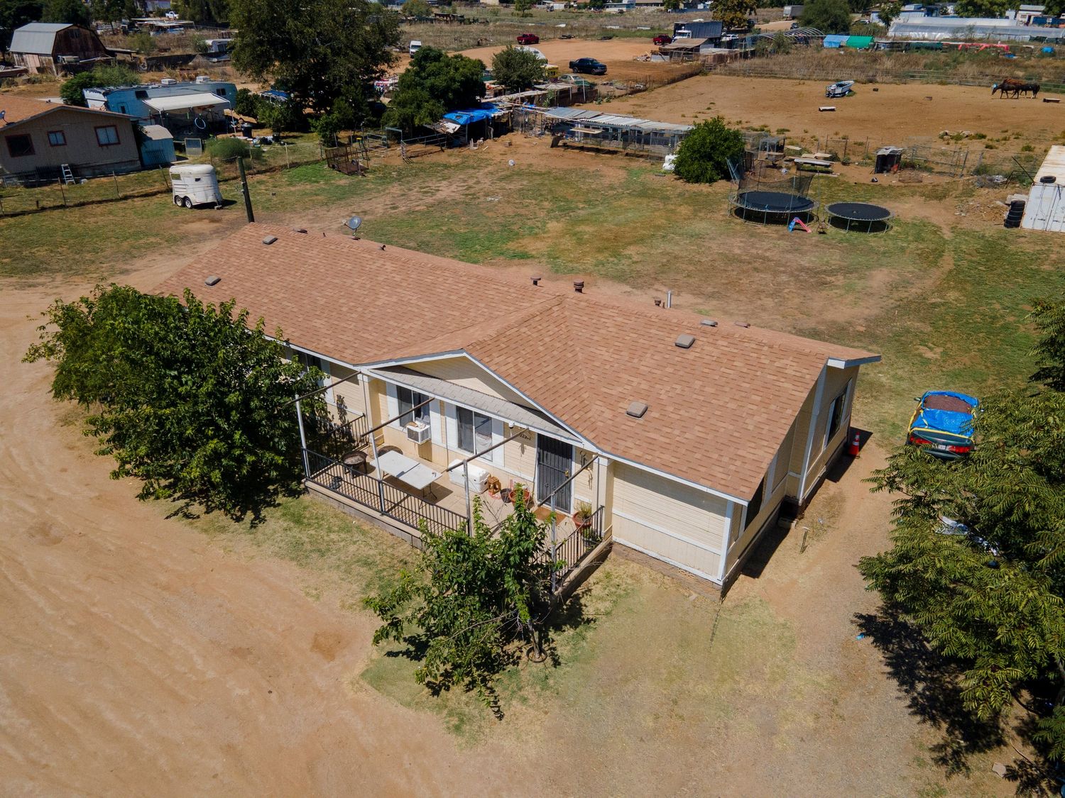 Aerial view of single-story ranch home on dry rural lot with mature trees and open land surroundings.