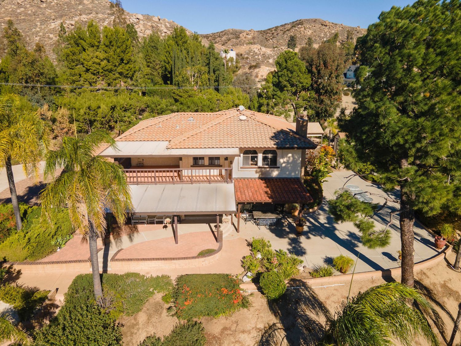 Aerial view of a single-story home with a tile roof, balcony, lush trees, and a hillside backdrop in California.