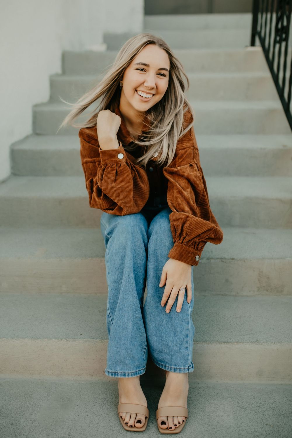 A relaxed portrait on concrete steps wearing a brown jacket and jeans.
