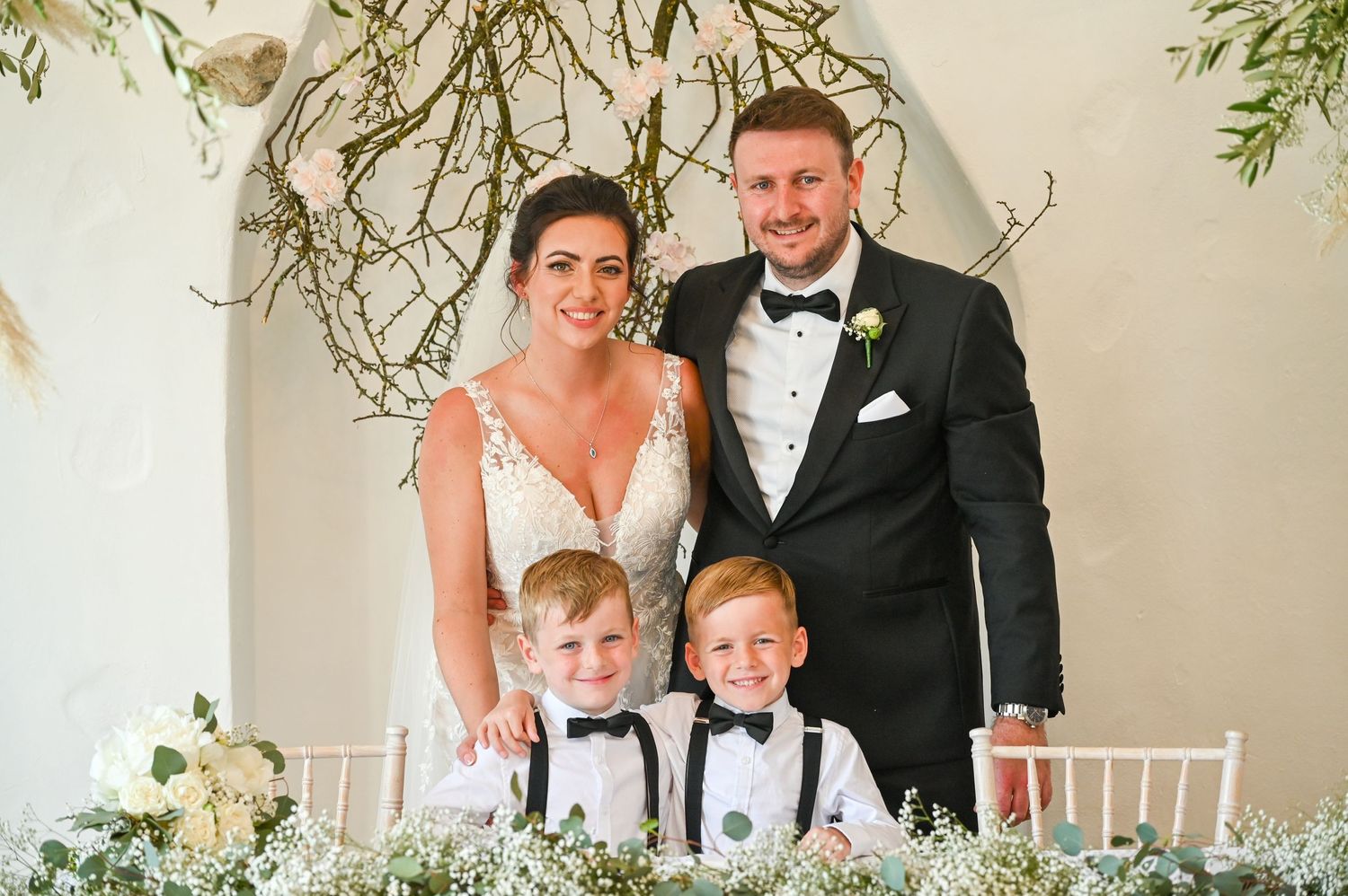 A wedding portrait with two young children in formal attire under a branch-decorated arch.
