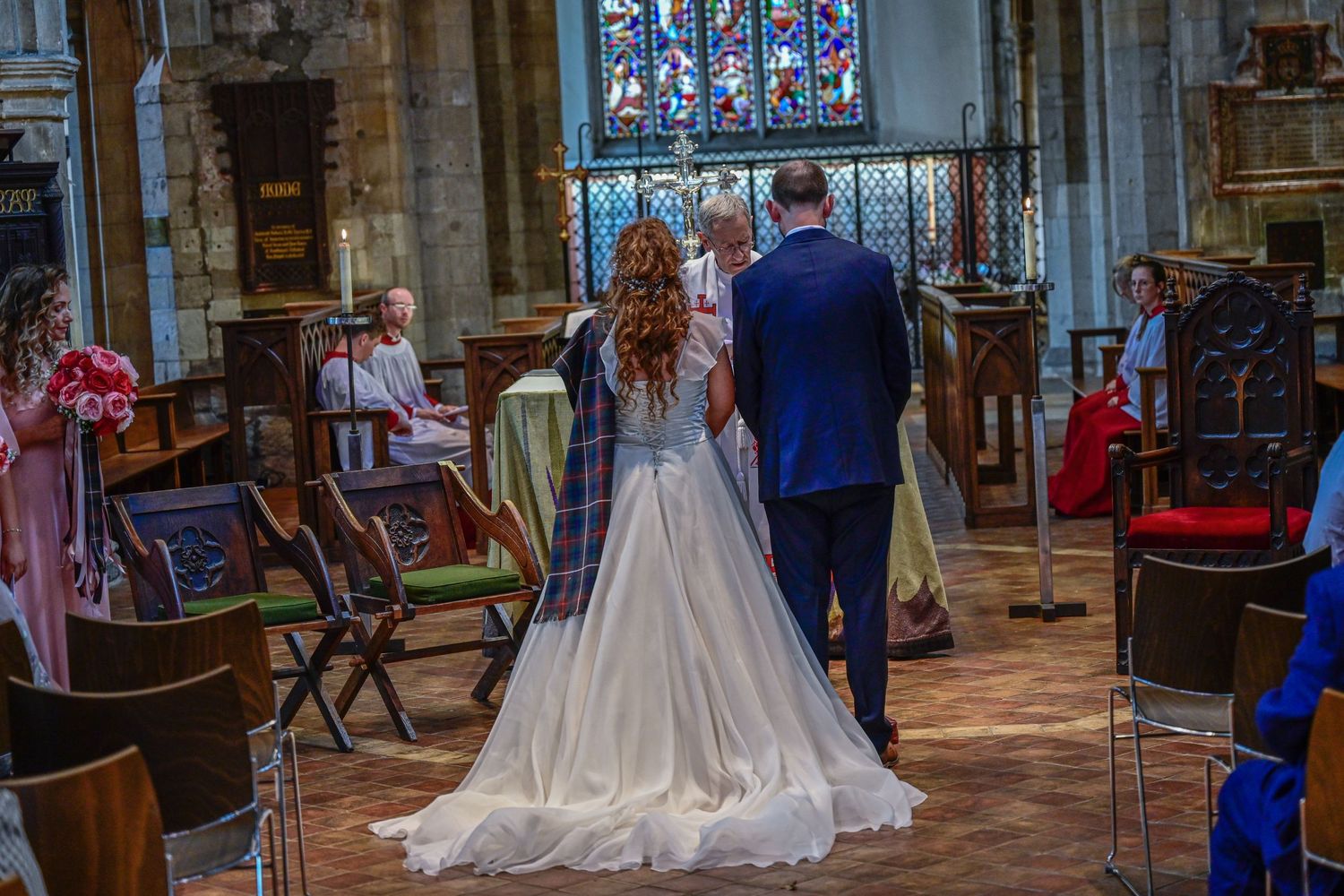 Back view of wedding couple standing at church altar with ornate stained glass window in background.