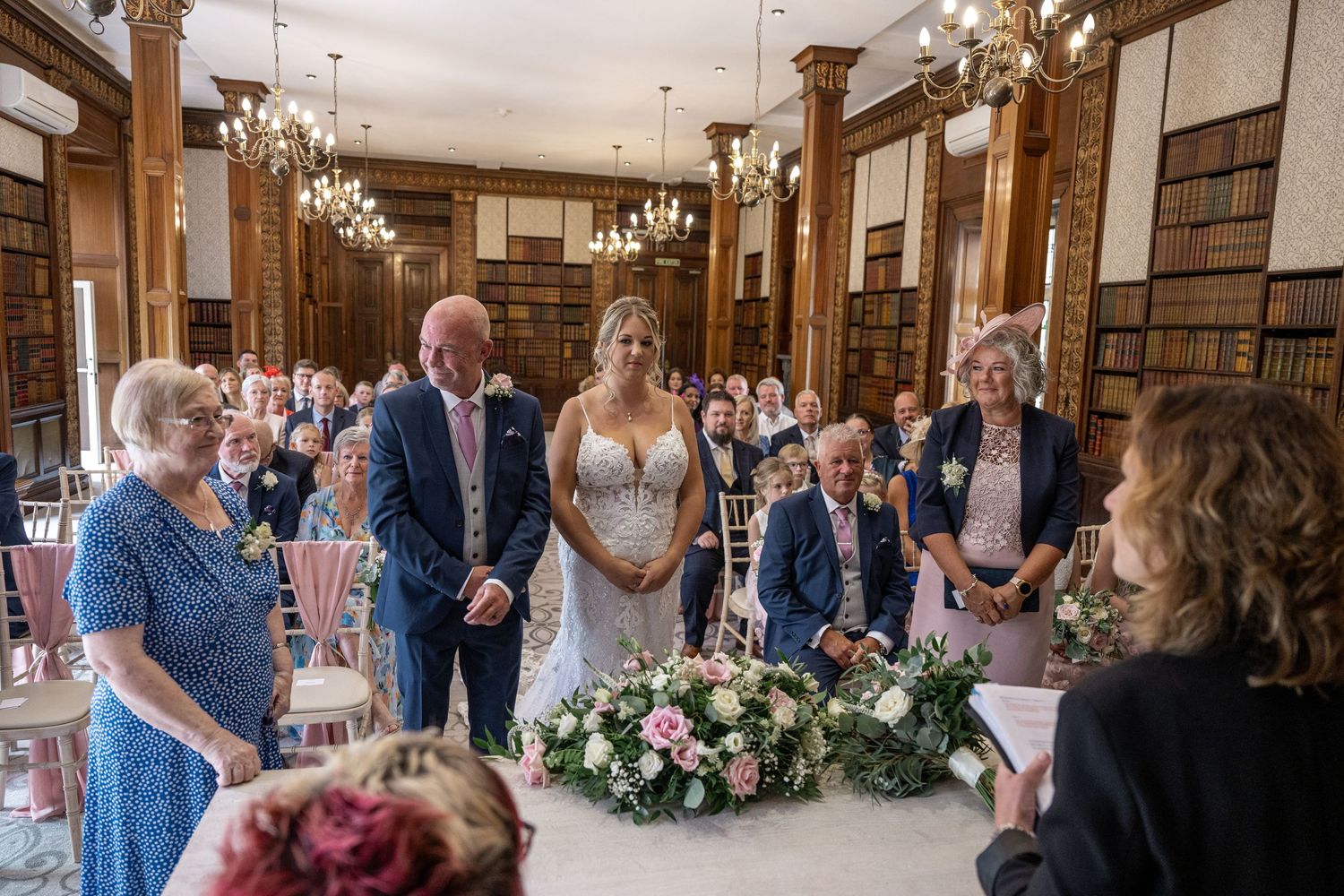 Wedding party gathered around a decorated table during ceremony in historic library setting.