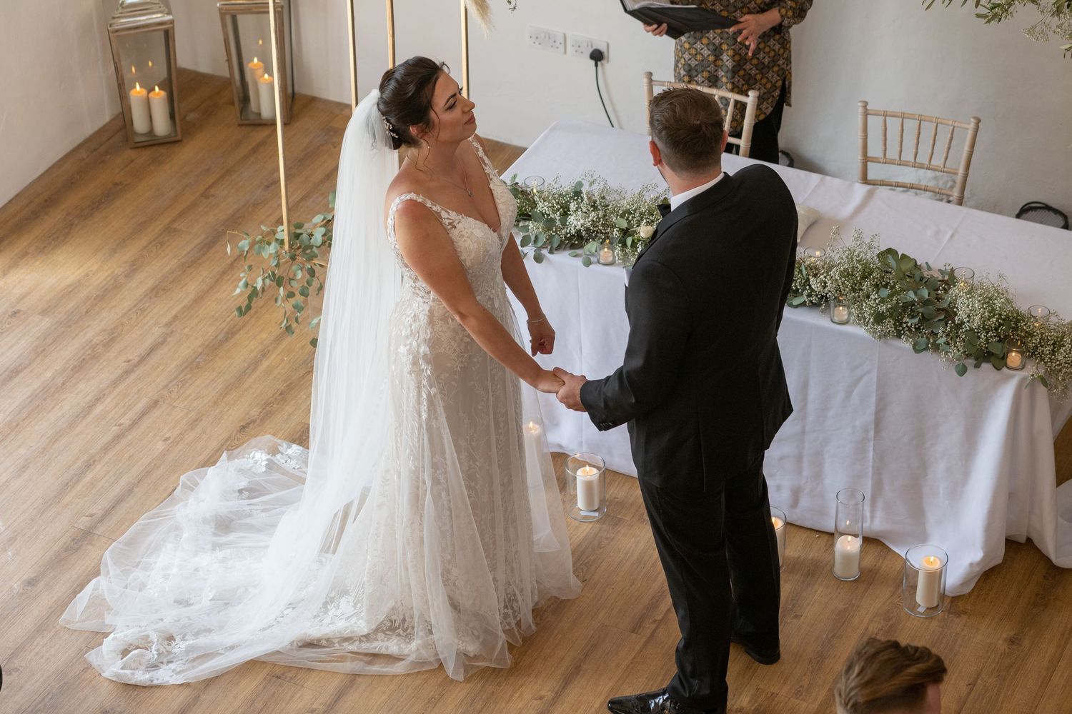 A wedding ceremony takes place in a bright room with white floral decorations and wooden floors.