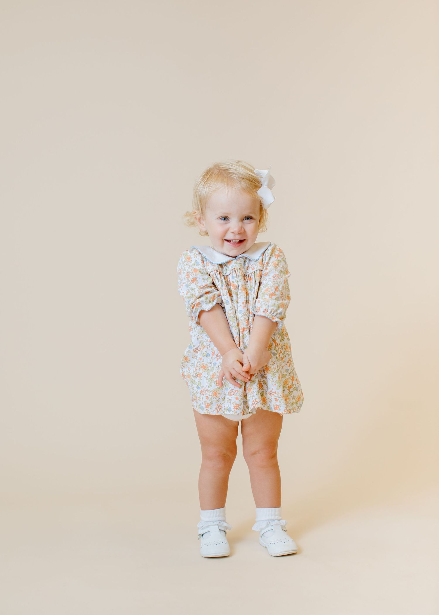 Toddler in floral dress and white shoes smiling shyly at camera.