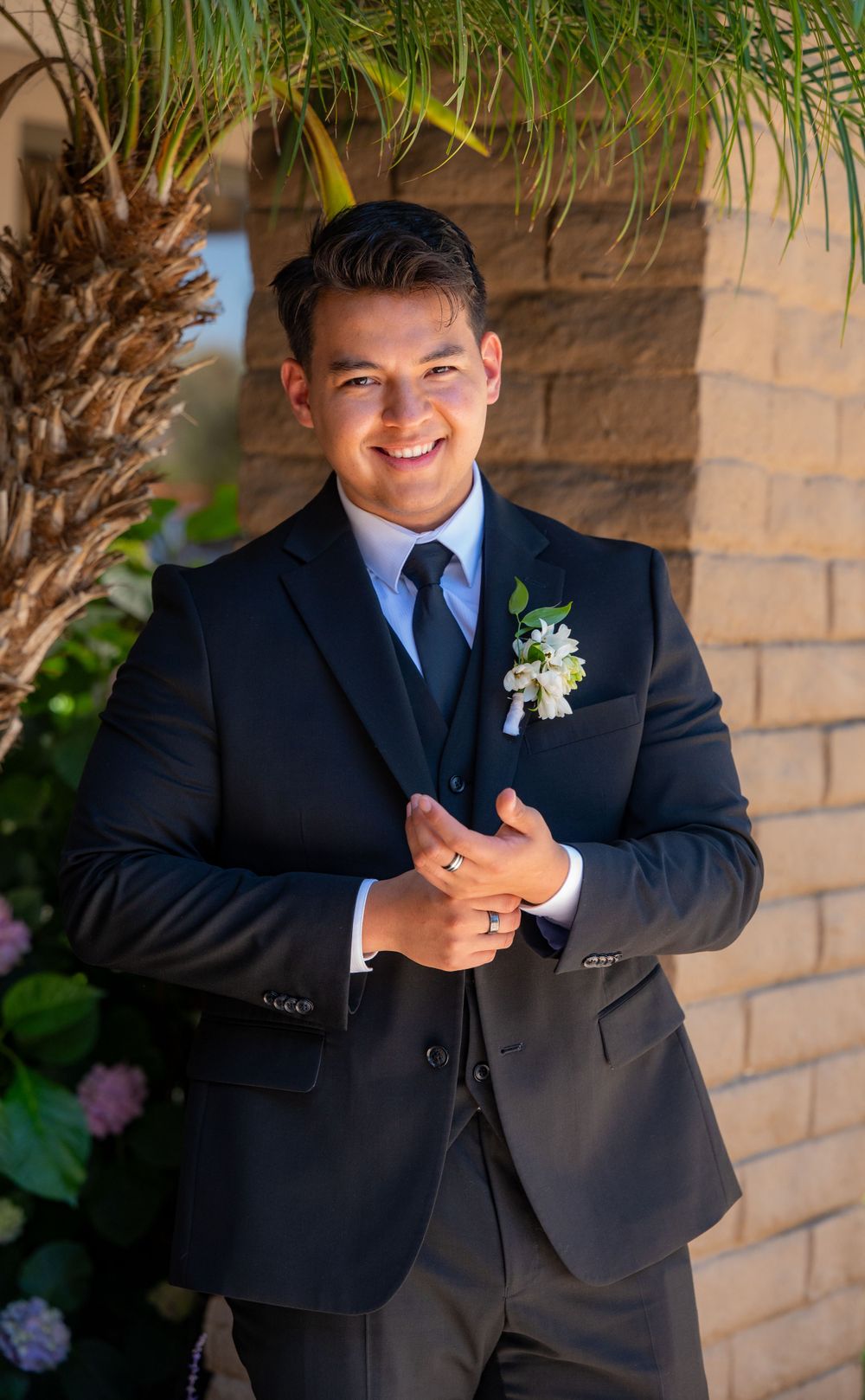 Groom in navy suit with boutonniere poses outdoors near palm trees.
