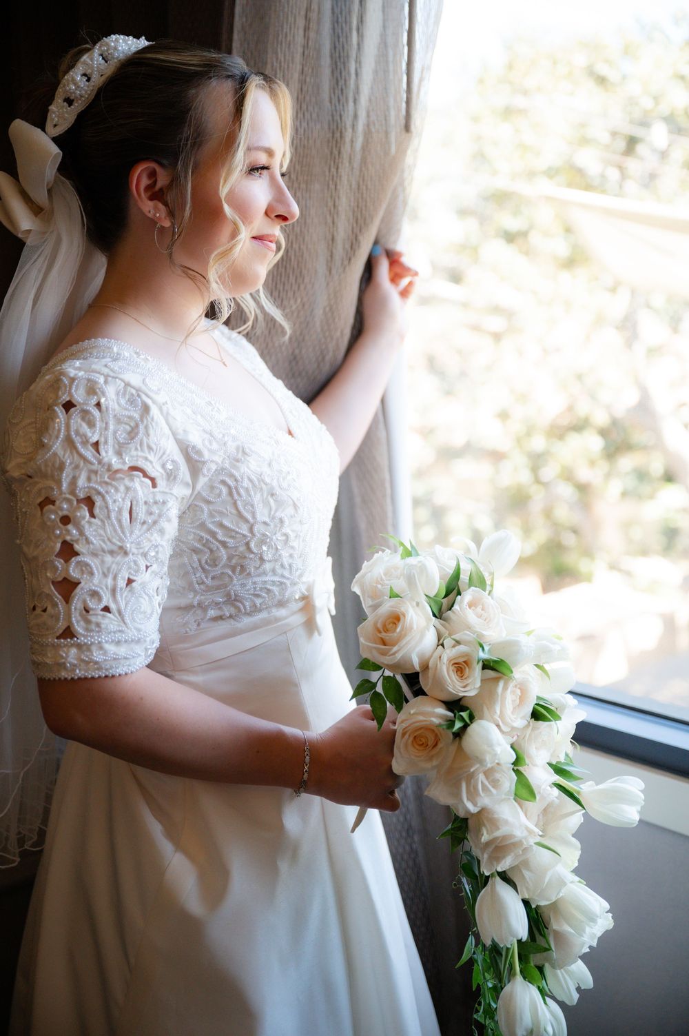 Bride in lace sleeve wedding dress holds bouquet of cream roses while looking out window.