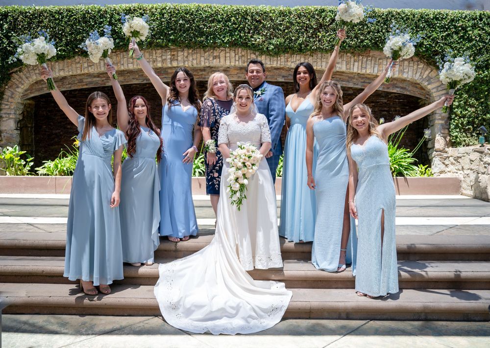 Bridal party in powder blue dresses poses with bride in white gown under an ivy-covered archway.