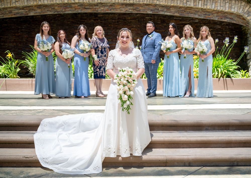 Bride in white gown with cascading train poses on steps with wedding party in blue dresses.