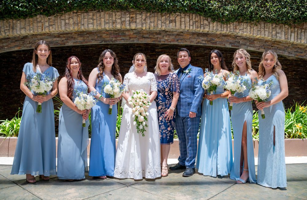 Wedding party wearing light blue dresses and formal attire gather for a group photo under an arch.