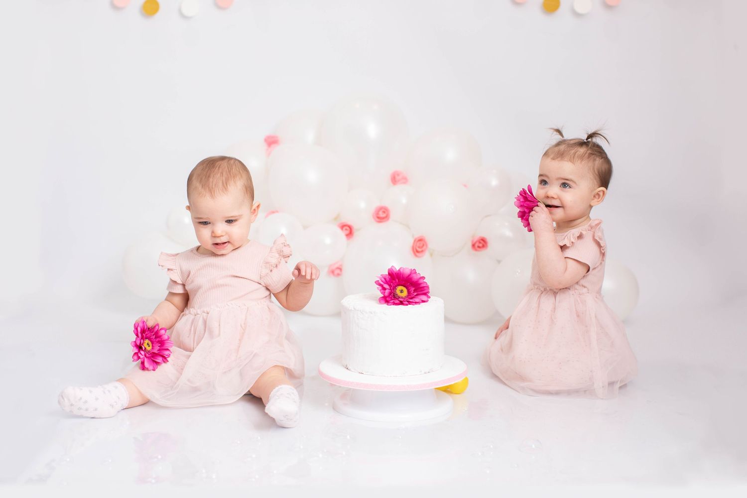 Twin baby girls in pink dresses with pink flowers celebrating their first birthday with a white cake.