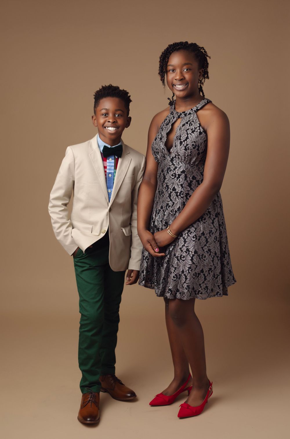 Fashionable duo poses in complementary outfits with a beige blazer and patterned dress against brown background.