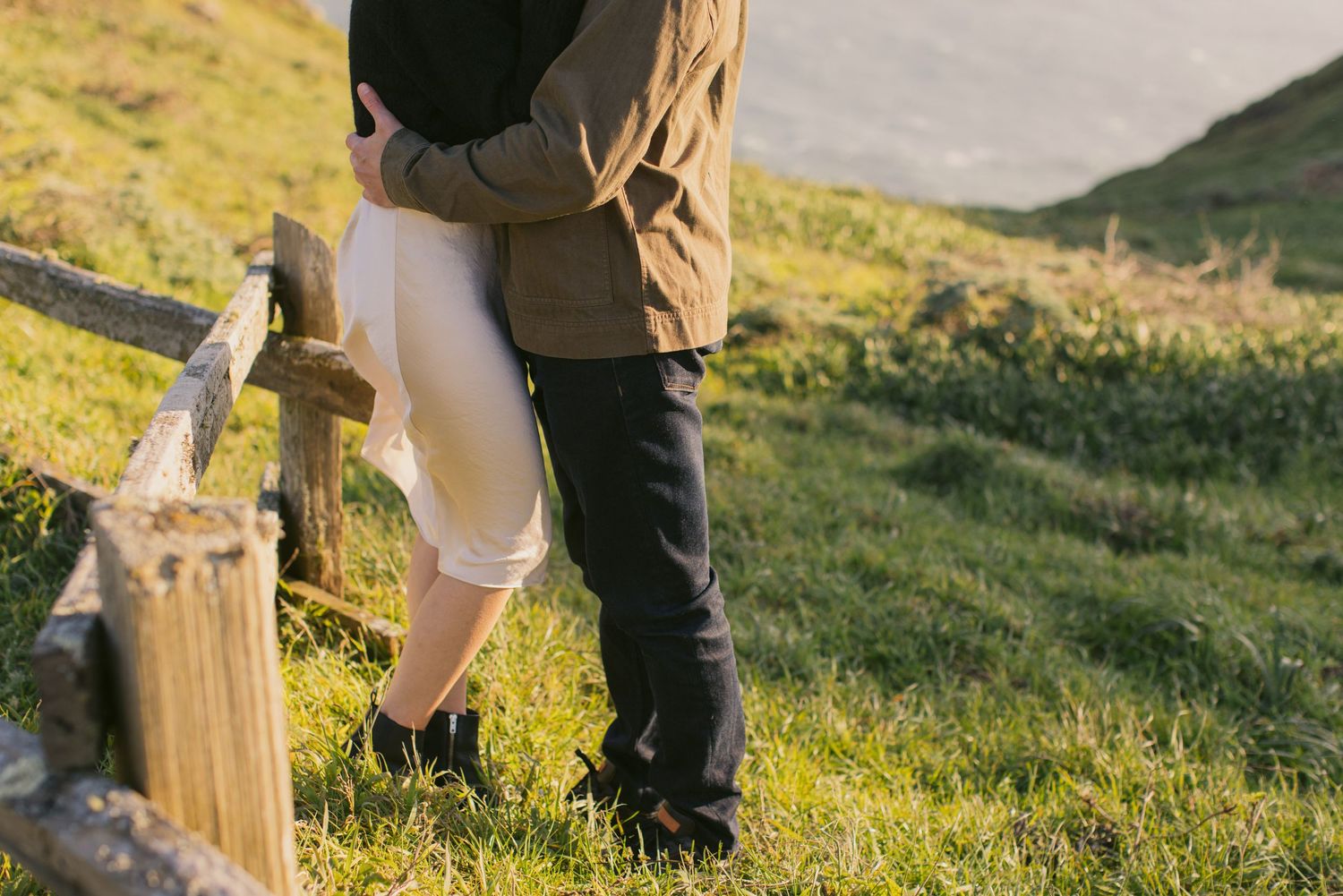 Close-up of a couple's legs standing by a wooden fence post on a grassy hillside.