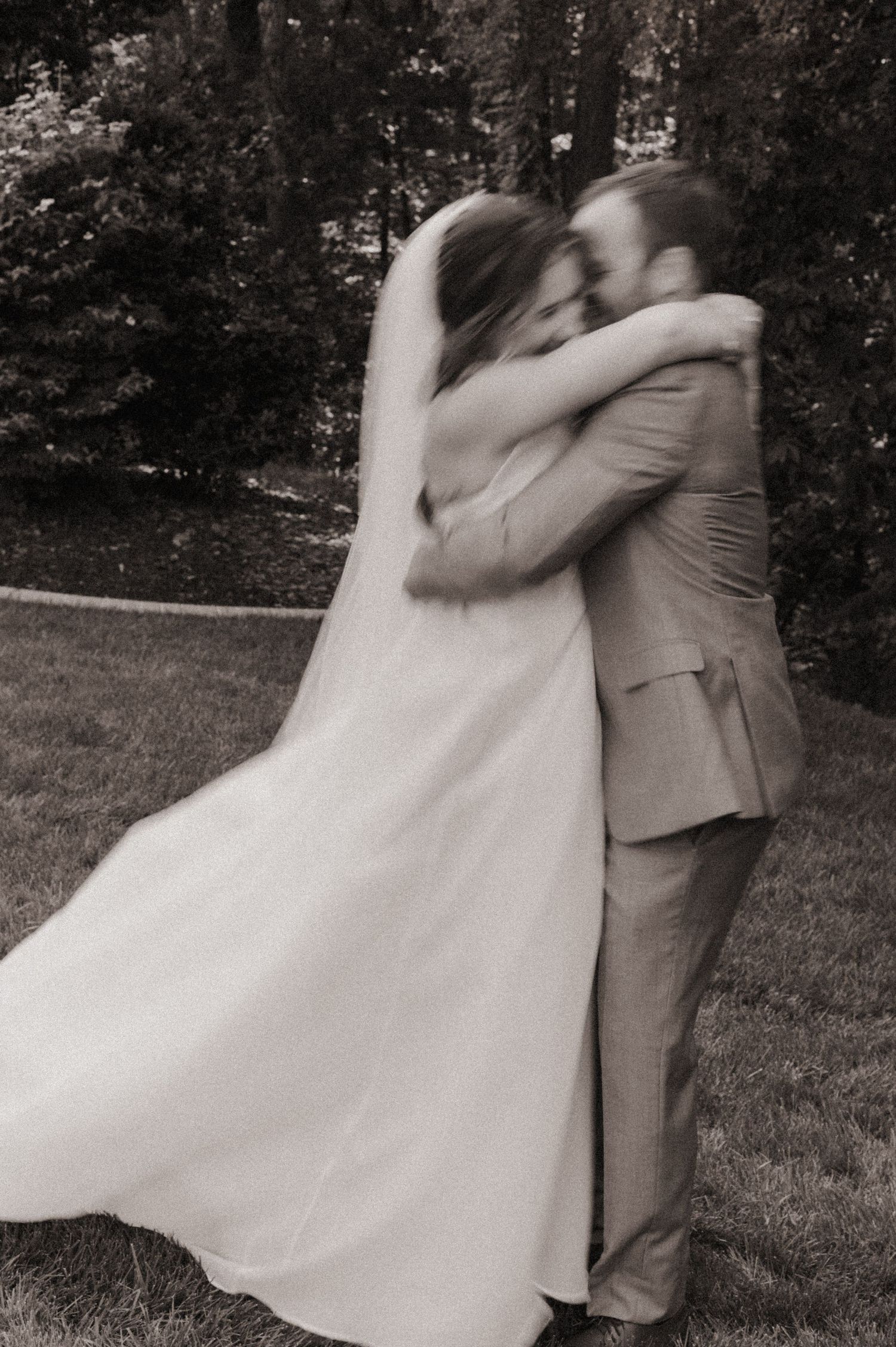 A black and white photo capturing an intimate wedding embrace outdoors with the bride's veil flowing in the wind.