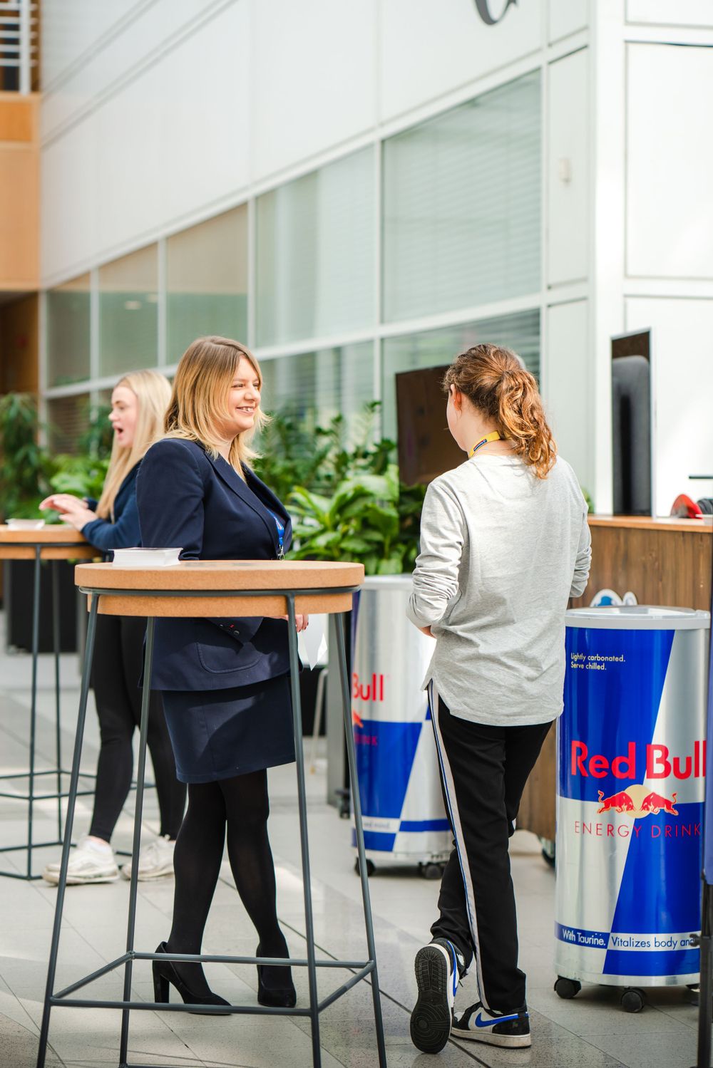 People socializing at high tables near Red Bull coolers in office setting.
