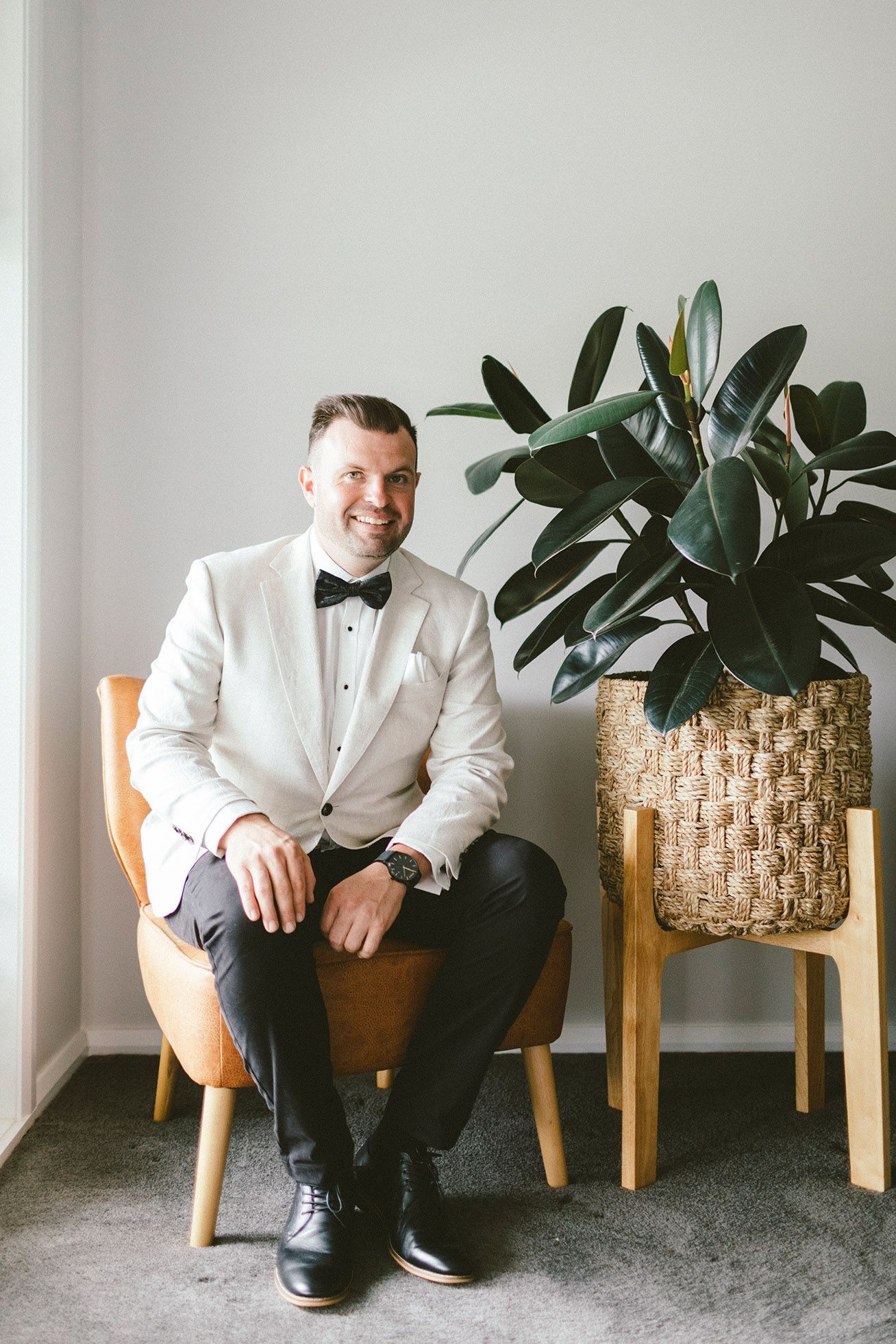 A person wearing a white tuxedo jacket and black bow tie seated in a wooden chair.