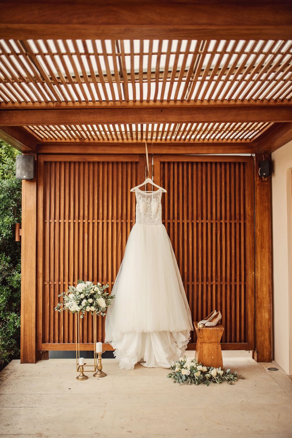 White wedding dress hanging on wooden pergola with floral arrangements and shoes below.