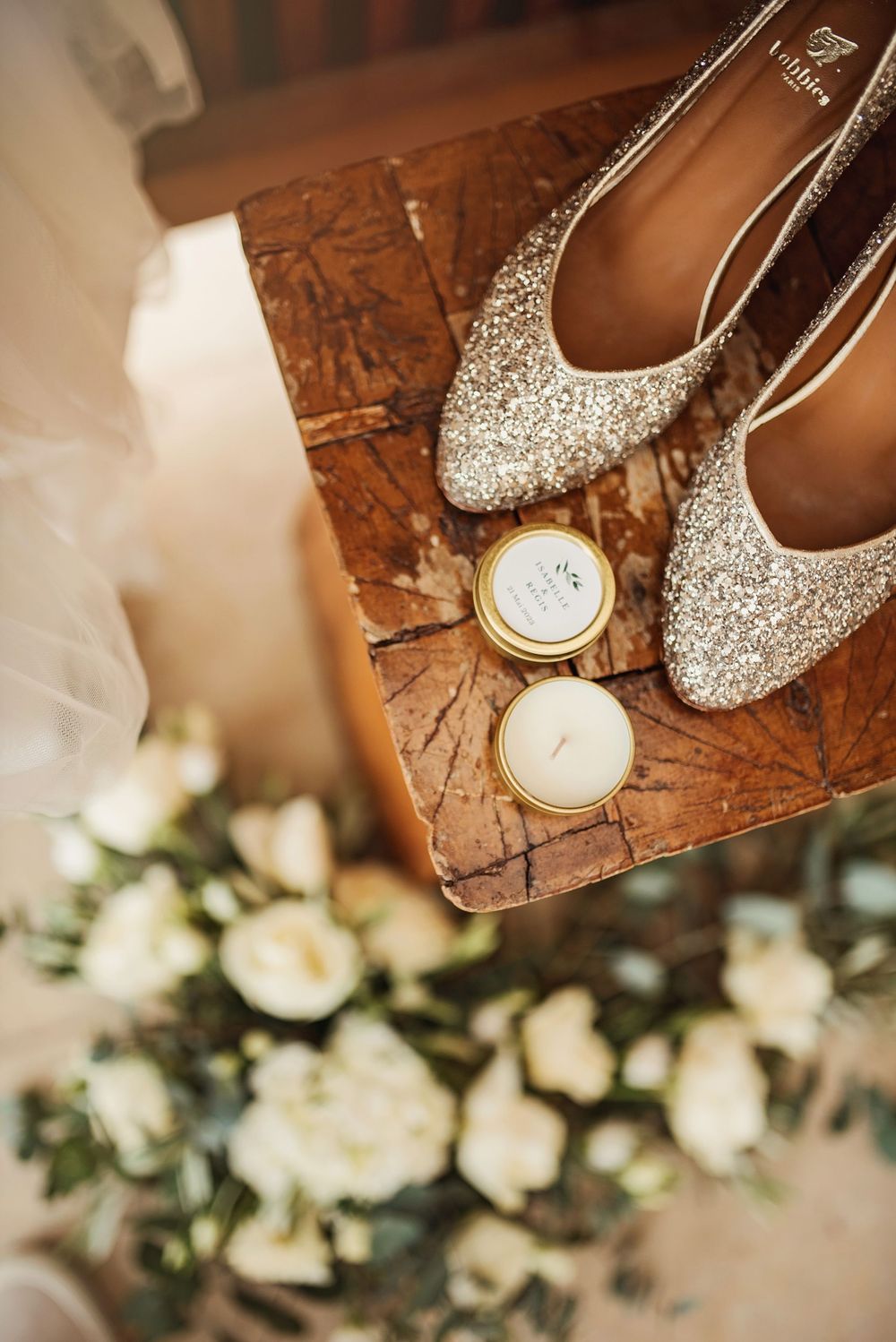 Glittery bridal shoes and candles on wooden surface with white roses in background.
