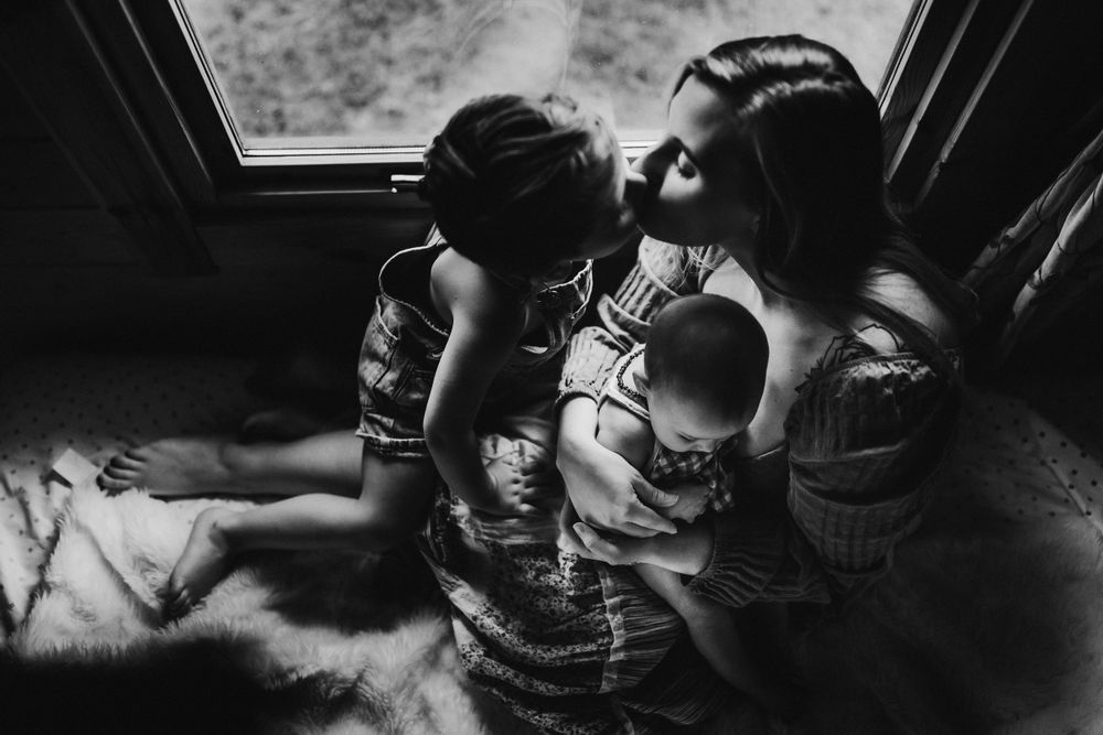 Black and white photograph of a tender family moment beside a window in natural light.