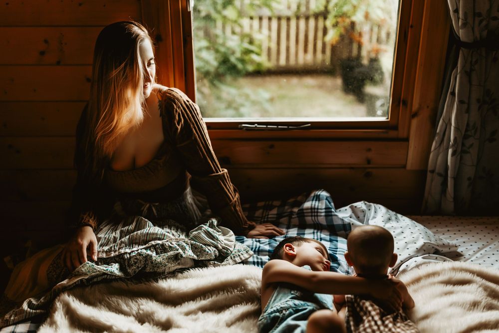 Person sitting by window with child on bed in morning sunlight.